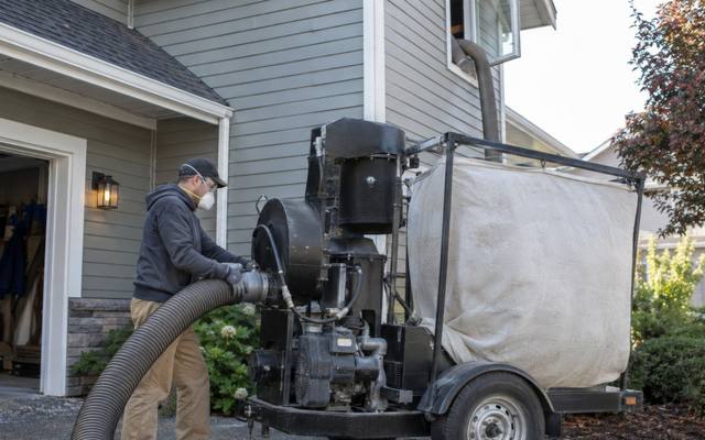 Trailer-mounted insulation removal vacuum with a big collection bag in a driveway and a hose running to the house