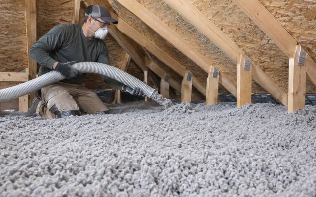 Worker holding a hose over a deep, even layer of grey loose-fill insulation covering an attic floor