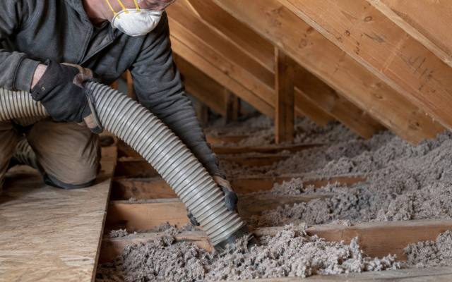 Worker using a vacuum hose to pull grey loose-fill from between attic joists during insulation removal